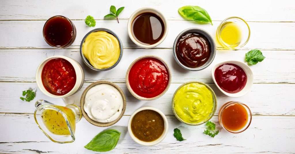 a bunch of condiments in little bowls on top of a whitewashed table. Mustard, ketchup, bbq sauce, tartar sauce, olive oil, salad dressing. top 10 condiments dogs should not eat.