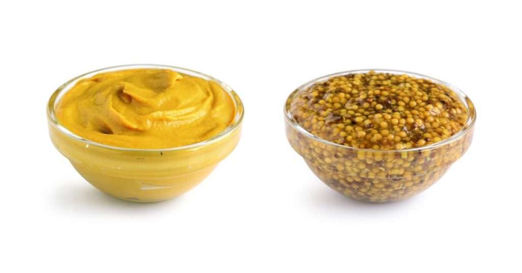 two small glass bowls with mustard in them, the one on the right has mustard with seeds in it, the left bowl is just yellow mustard
