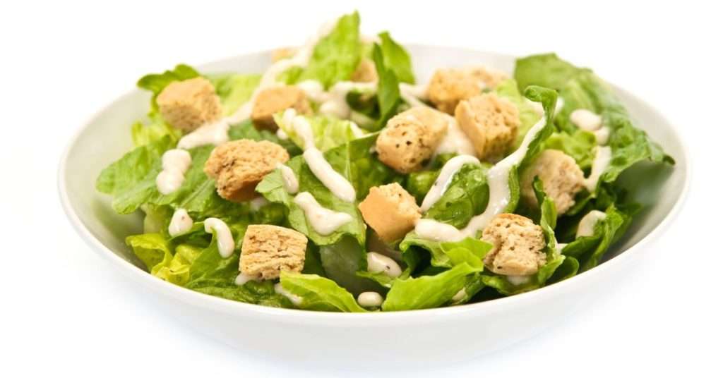 Dogs should not eat salad dressing.  Caesar salad dressing is not safe for dogs. Caesar salad in a white bowl with croutons on top.  The dressing is thick and on top of the lettuce.